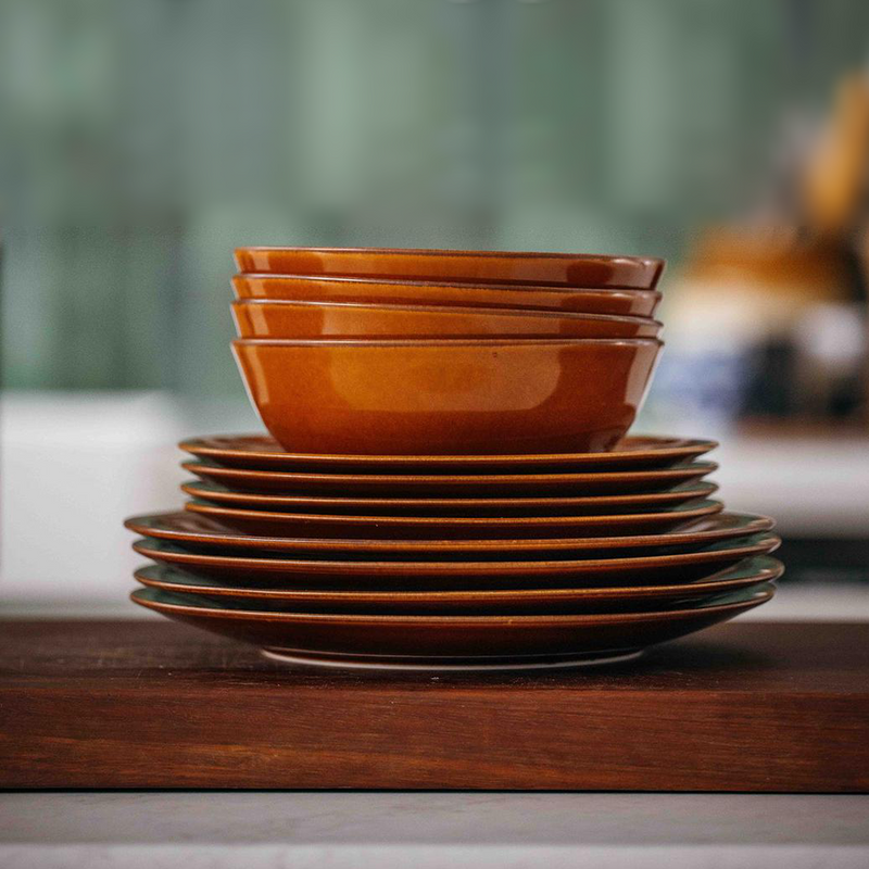 Adam Liaw Everyday Noritake 12-piece dinnerware set in Aemyu warm amber reactive glaze, stacked as four large plates, four medium plates, and four bowls on a wooden surface
