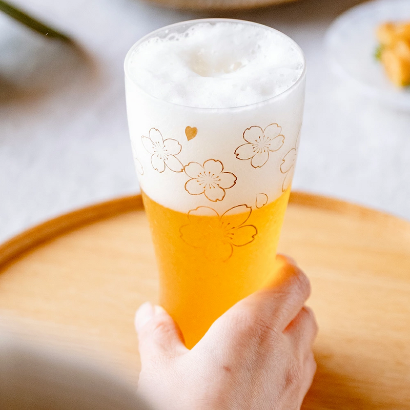 Hand holding the beer glass filled with frothy beer, highlighting the golden floral pattern.

