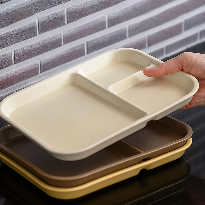 Close-up view of a beige Aito Mino Yaki divided plate with a hand reaching to open its lid, showcasing its compartment. The plate is placed against a brick background.