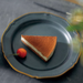 A matte black ceramic plate with scalloped edges, accented by a delicate gold trim, showcasing a single slice of tiramisu and a red cherry.