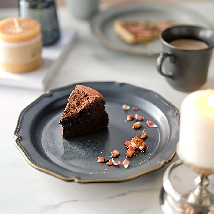 A setting featuring the same black ceramic plate with golden edging, holding a rich piece of chocolate cake, paired with a matching black mug on a white tabletop.