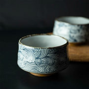 An Arahami Japanese matcha bowl with an intricate blue 'wild wave' pattern, in focus on a dark surface with a second bowl blurred in the background.