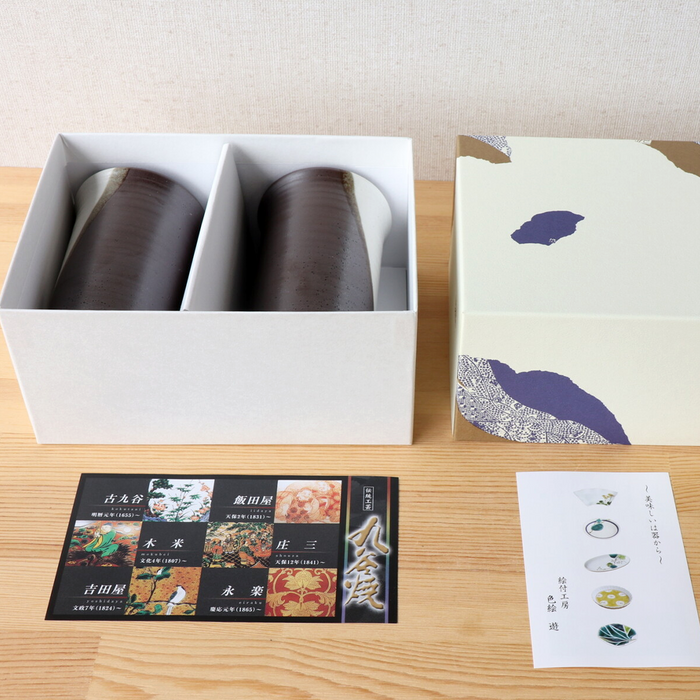 A neatly arranged gift box containing two Kutani ware tumblers. The tumblers are nestled in individual compartments within a white box, which is partially covered by a lid featuring traditional Japanese patterns in soft beige and blue tones. Additional cards or inserts with Japanese text and designs are displayed alongside the box.