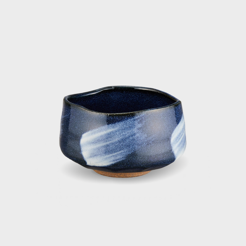 A studio product shot of the blue glaze matcha bowl with a white brushstroke, showing its irregular shape and unglazed clay foot. 