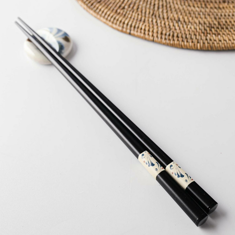A pair of black Japanese chopsticks with a decorative white band featuring a blue Karakusa (arabesque) pattern, resting on a ceramic chopstick rest. A woven placemat is visible in the background.
