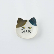 White Japanese cat cutlery rest with multicoloured ears on a textured ceramic plate.