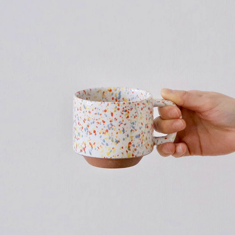 A hand holding a white CHIPS Stackable Mug with colourful orange, yellow, and blue splashes, featuring a rustic unglazed clay base.