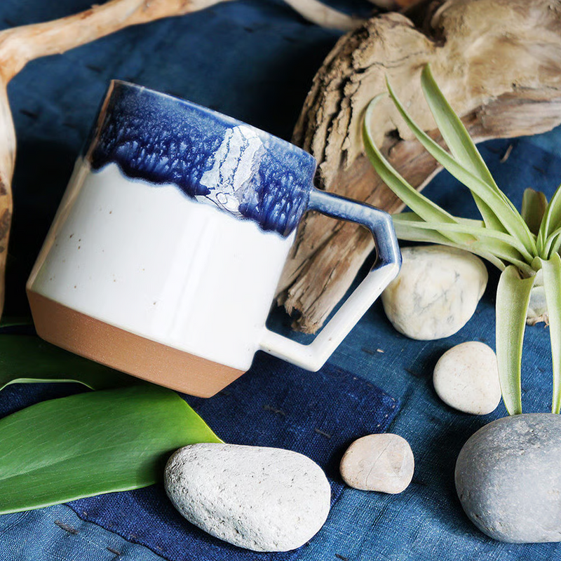 A styled lifestyle photo of the CHIPS Stack Mug in White Navy Drop, placed among driftwood, stones, and a plant on a blue fabric background.