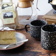 Two black CHIPS splatter mugs on a wooden table with a slice of cake and coffee-making equipment.