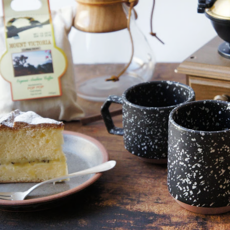Two black CHIPS splatter mugs on a wooden table with a slice of cake and coffee-making equipment.