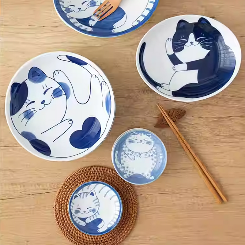 Four Daitoua Neko Chigura oval serving bowls with hand-painted blue and white cat designs, each featuring a different playful cat illustration. The bowls are displayed on a light wooden surface, with two of them adorned with small flowers.