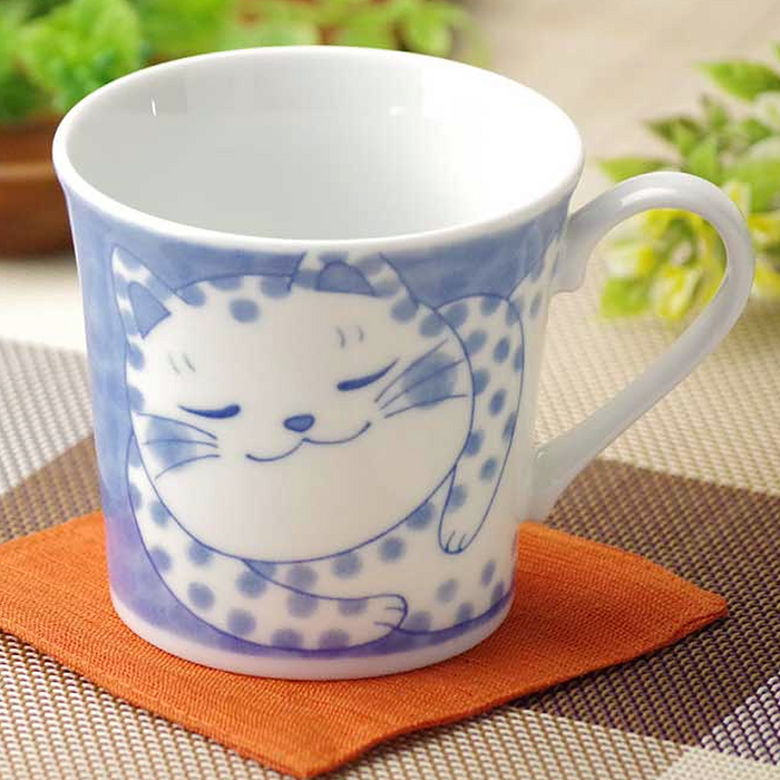 Artistic blue cat design on a white ceramic mug placed on an orange cloth with a blurred greenery background.