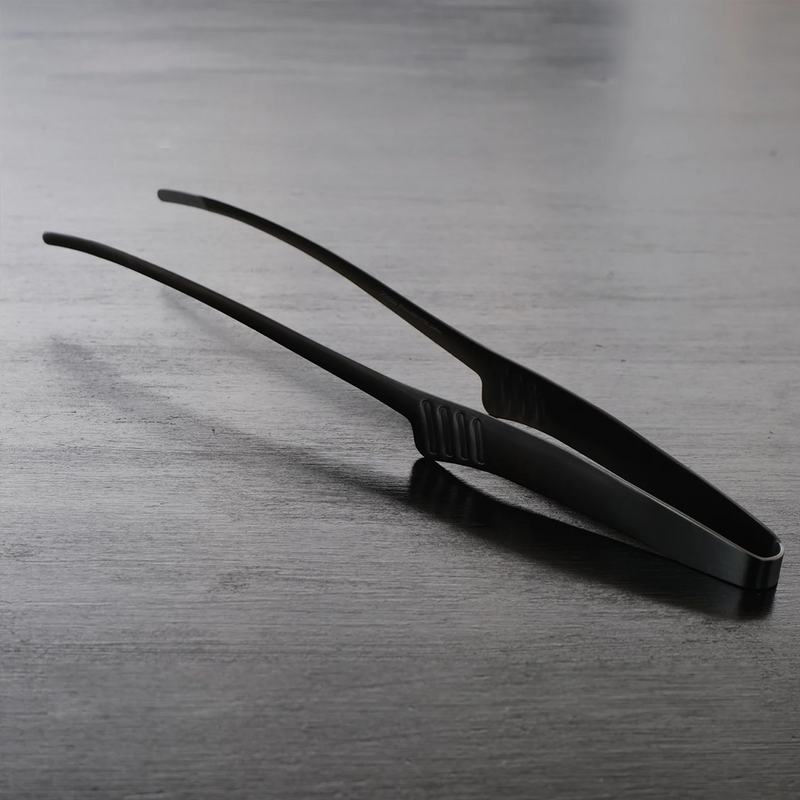 EBM matte black tongs photographed on a dark wood surface highlighting the sleek finish and fine tips.


