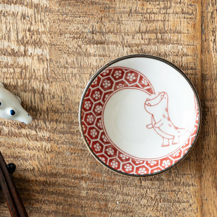 A red-patterned mini plate featuring a playful dinosaur illustration, set on a textured wooden table, highlighting its artisanal details.

