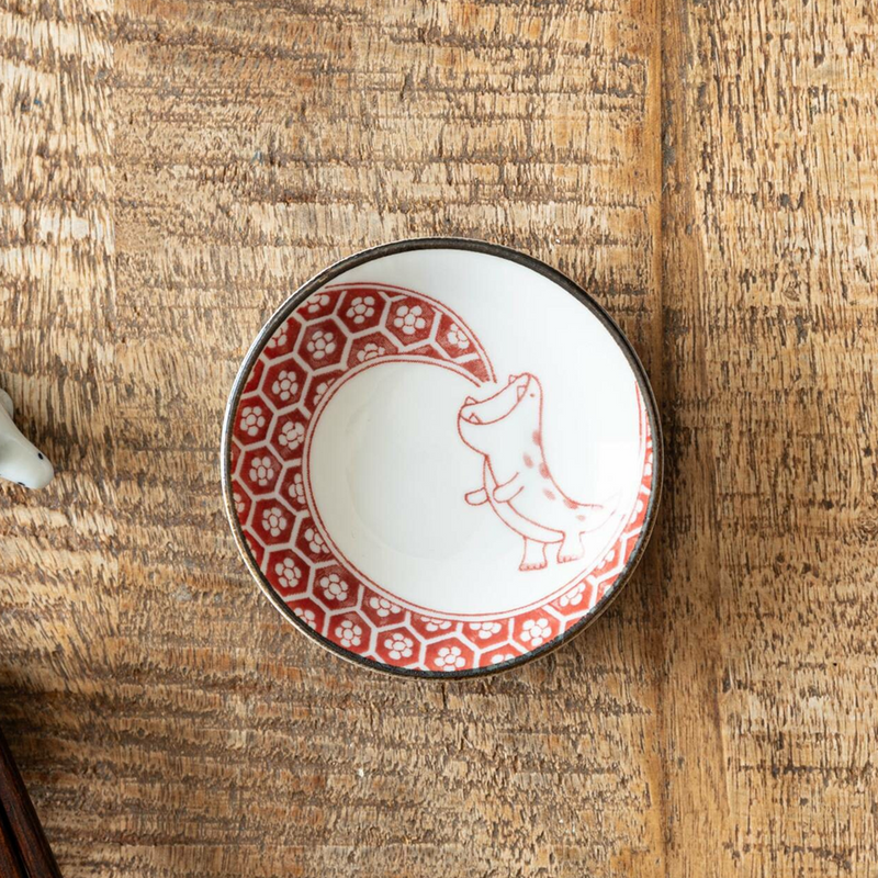 A red-patterned mini plate featuring a playful dinosaur illustration, set on a textured wooden table, highlighting its artisanal details.