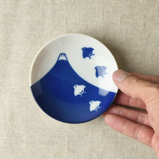 A small round porcelain side plate in a hand, displaying a vibrant indigo Mount Fuji design with plover birds on a white background over a neutral fabric backdrop.






