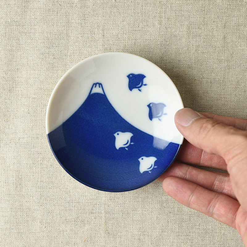 A small round porcelain side plate in a hand, displaying a vibrant indigo Mount Fuji design with plover birds on a white background over a neutral fabric backdrop.






