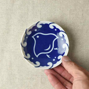 A hand holding a small, round blue and white ceramic plate with a bird design on a beige fabric surface. In the background, a stack of similar plates features intricate blue patterns of birds and waves, characteristic of traditional Japanese porcelain.