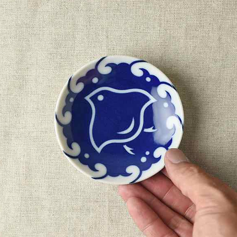 A hand holding a small, round blue and white ceramic plate with a bird design on a beige fabric surface. In the background, a stack of similar plates features intricate blue patterns of birds and waves, characteristic of traditional Japanese porcelain.
