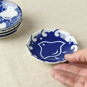 A hand holding a compact porcelain plate with a bold indigo bird design against white, with additional plates stacked in the background

