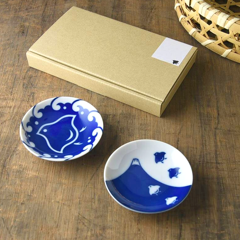Two small round porcelain side plates with bright indigo designs (Mount Fuji and stylised plover birds), shown beside a rectangular kraft box on a wooden table
