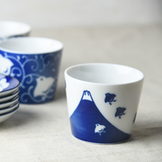 A white ceramic cup with a blue Mount Fuji and bird pattern depicted around the lower portion, set on a neutral-toned background.
