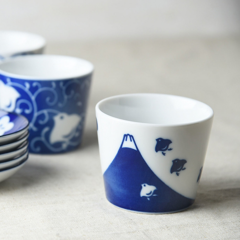 A white ceramic cup with a blue Mount Fuji and bird pattern depicted around the lower portion, set on a neutral-toned background.