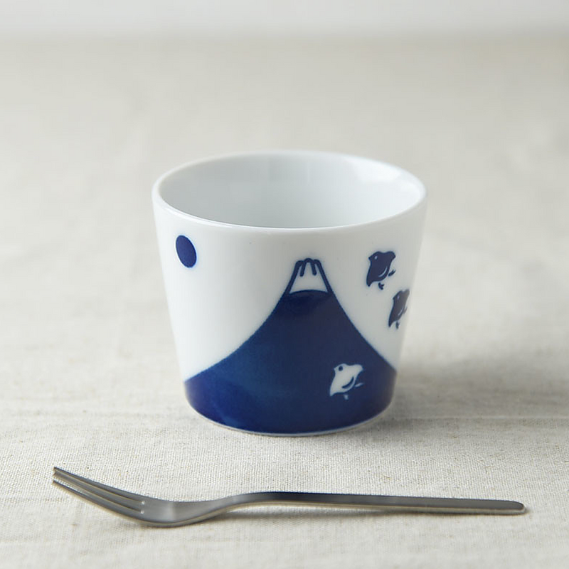 A white ceramic cup on a linen surface, displaying a vivid blue Mount Fuji and bird pattern, next to a dark blue cup.