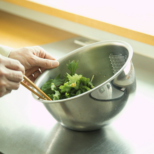 Fujii Stainless Steel 3-Way Rice Washing Bowl being tilted to drain water through its side perforations while holding leafy greens.