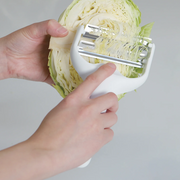 Hands holding a white Pearl Life double-blade cabbage peeler, shredding a green cabbage half to create fine strips.