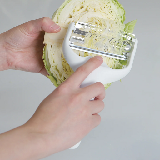 Hands holding a white Pearl Life double-blade cabbage peeler, shredding a green cabbage half to create fine strips.