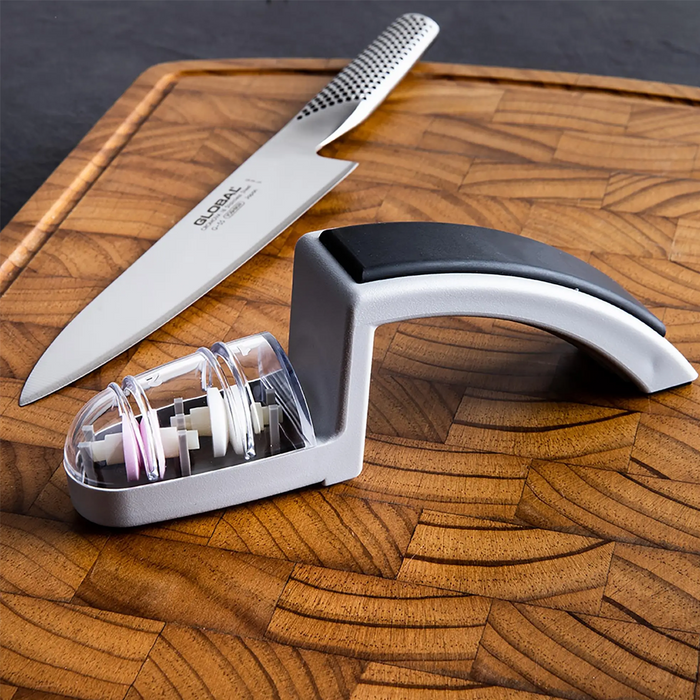 Side view of Global MinoSharp 2-stage ceramic sharpener with a curved silver handle and clear compartment housing pink and white sharpening wheels, on a plain background.