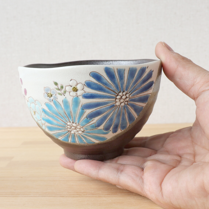 A close-up of a hand holding a blue Hana Floral Kutani bowl, emphasizing its intricate hand-painted design. The bowl features large, vibrant blue flowers with smaller white blossoms against a textured speckled grey base, showcasing its unique shape and artisanal craftsmanship.