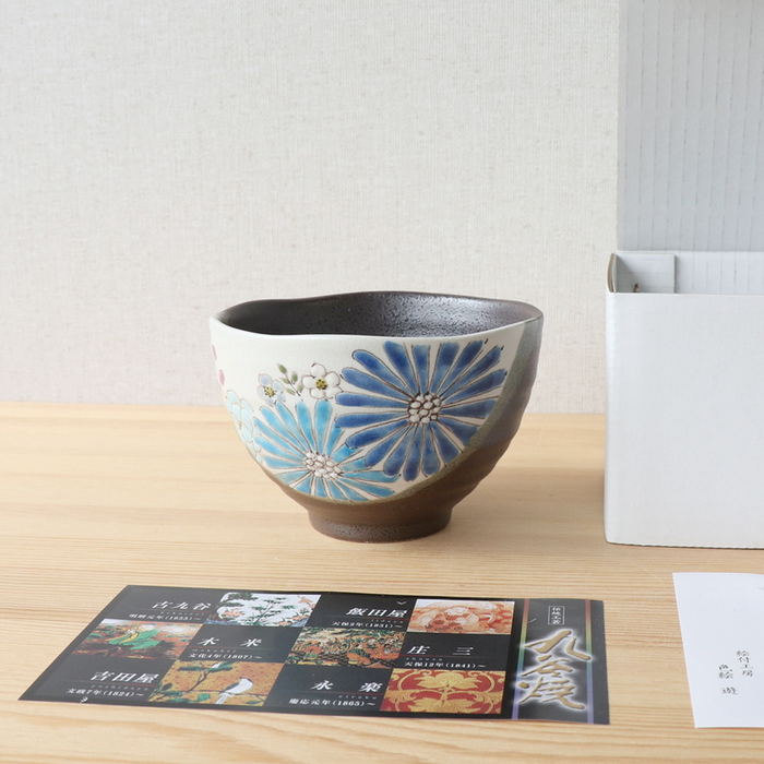 A blue Hana Floral Kutani bowl displayed on a wooden surface next to its packaging. The bowl features large, hand-painted blue flowers with smaller white and pastel-colored blossoms on a speckled grey base. The box and informational cards are visible in the background, emphasizing the bowl's high-quality presentation.