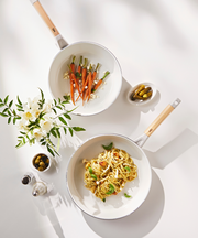 Styled top-down image of two Happycall Sonora ceramic frying pans with food — baby carrots in one, pesto pasta in the other — surrounded by olives and flowers.







