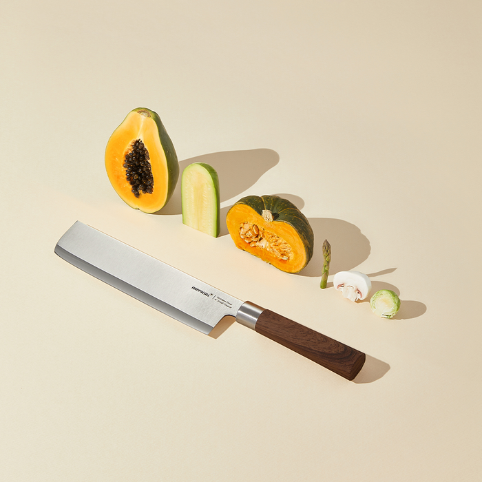 Happycall Woodique Nakiri knife with wooden handle displayed with cut pumpkin and other vegetables on a light background.