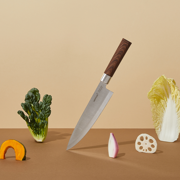 Happycall Woodique chef's knife with wooden handle standing upright surrounded by various vegetables including pumpkin, onion, and leafy greens.