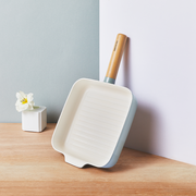 The square 22cm Happycall Zium ceramic nonstick induction grill pan, showing its sage green exterior, white ridged interior, and light wood handle, angled on a white background.