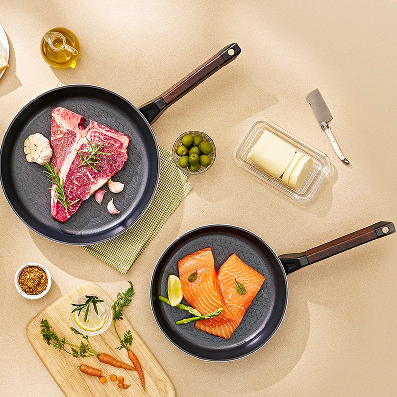 Happycall Royal Series 24cm and 28cm frypan set on a beige surface with salmon, steak, and cooking ingredients.