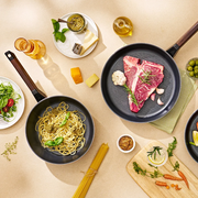 Top-down view of Happycall Royal series frypan and wok filled with steak and pasta dishes, surrounded by ingredients like tomatoes, pasta, and olive oil on a textured beige surface.