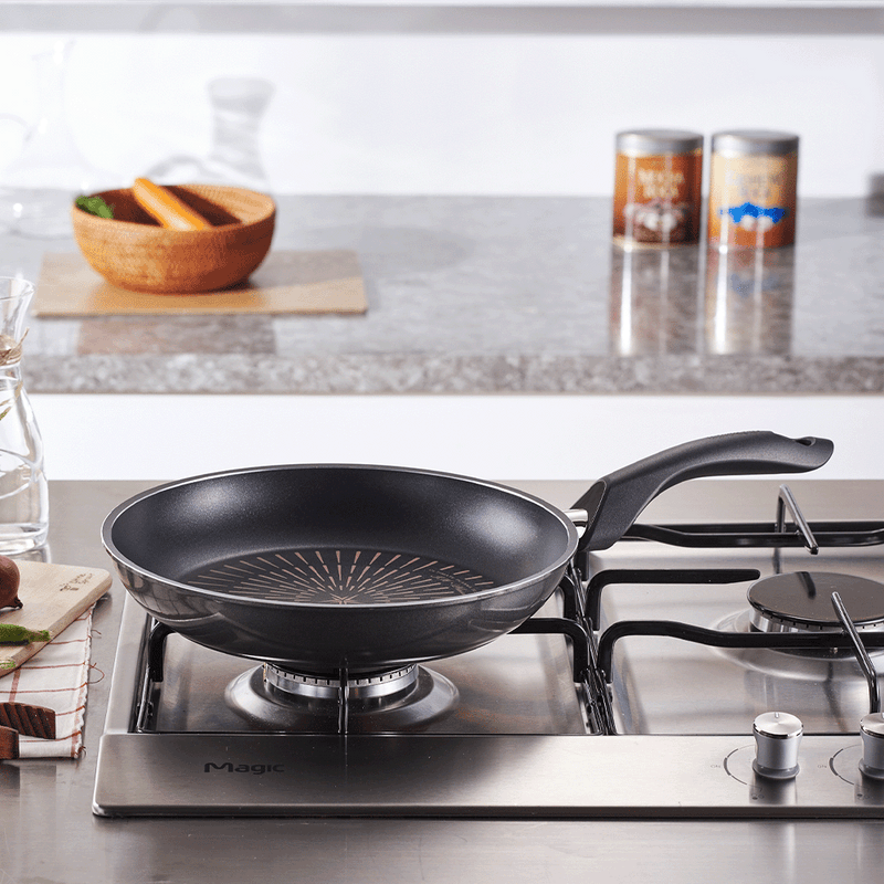 Happycall Plasma Titanium Frypan on Gas Stove: A black nonstick frypan with a circular pattern on the interior, placed on a gas stove in a modern kitchen setting. The handle is ergonomically designed for comfortable grip.

