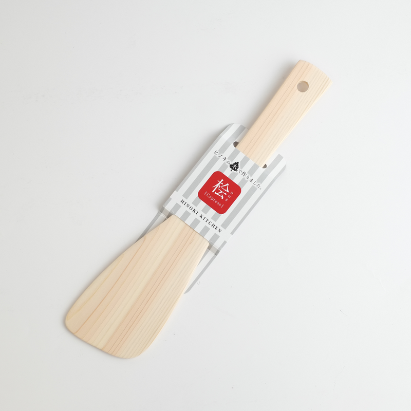 Hinoki Kitchen wooden spatula in natural Japanese cypress, showing fine wood grain and a wide angled head, with the branded paper band label reading "Hinoki Kitchen - Cypress" in a grey stripe design
