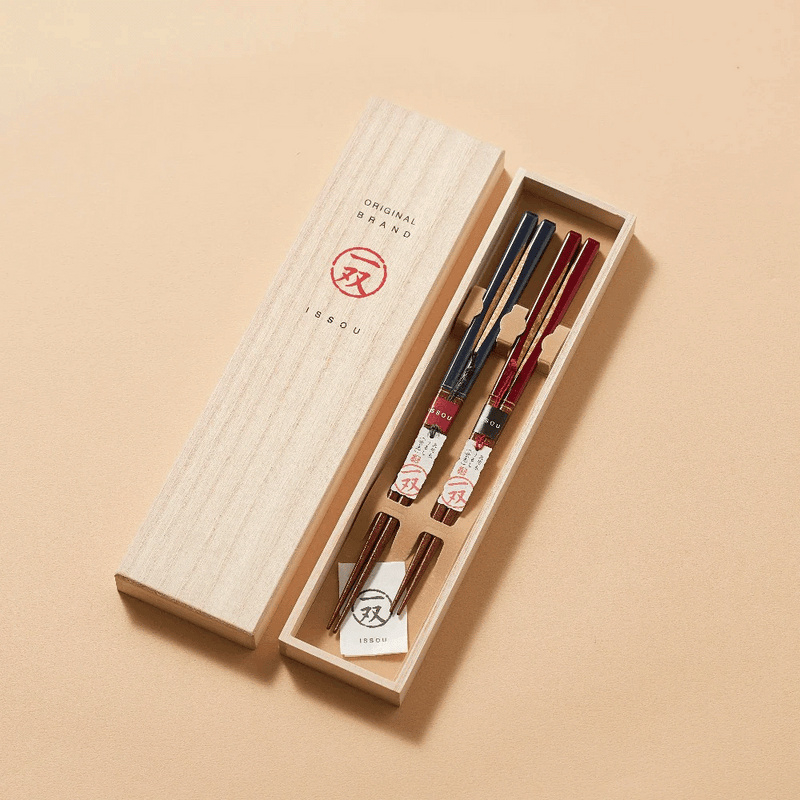 A set of Ishida Dream Droplets Wakasa Nuri Lacquerware Chopsticks in a Kiri wood box. The chopsticks have a natural wood color with a red and black lacquer finish and traditional Japanese patterns.
