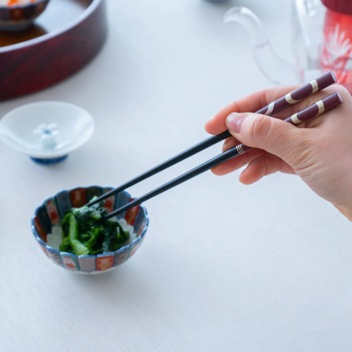 A hand holding a pair of burgundy Ishida Wakasa Nuri chopsticks, picking up food from a small ceramic dish during a meal.