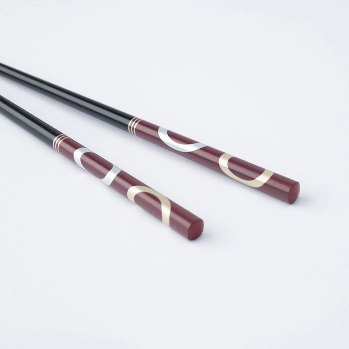 A pair of Ishida Wakasa Nuri chopsticks in burgundy with silver and gold ring patterns, laid out on a white background.