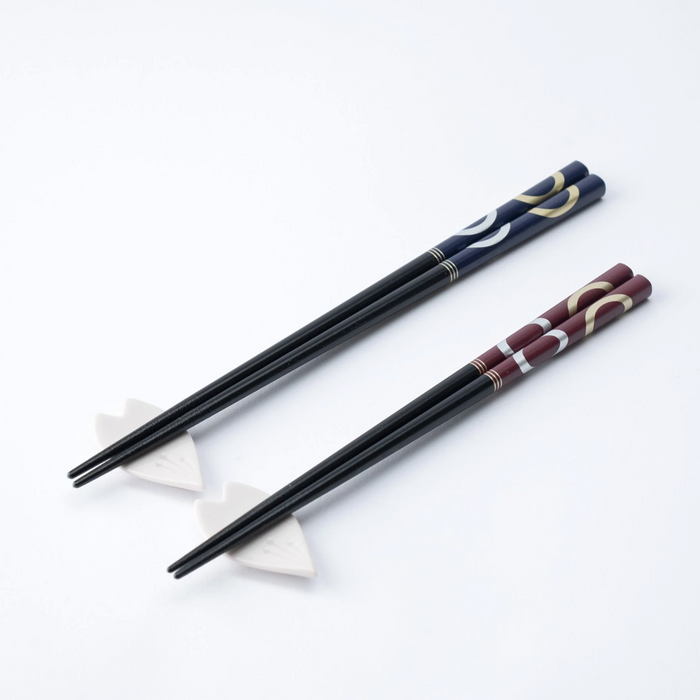 Two pairs of Ishida Wakasa Nuri lacquered chopsticks, one burgundy and one navy, resting on ceramic chopstick holders on a white surface.