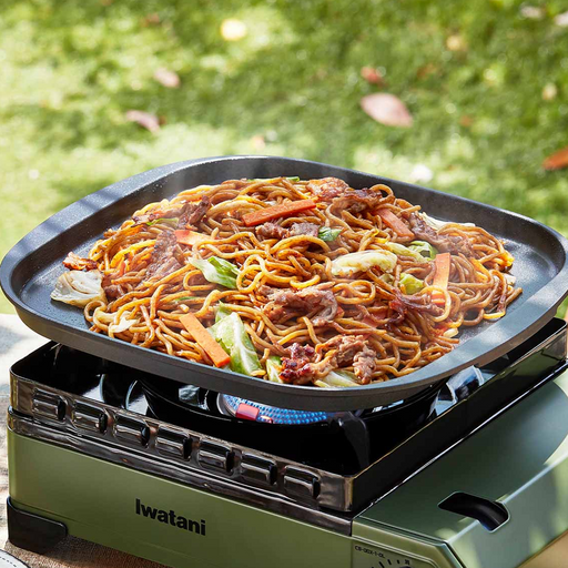 A 32cm Iwatani Teppanyaki Plate placed on a portable gas stove, used outdoors to cook a large portion of yakisoba noodles with vegetables and meat.