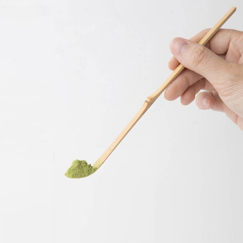 A hand holding a traditional Japanese bamboo matcha spoon (chashaku) with a scoop of green matcha powder on the tip.