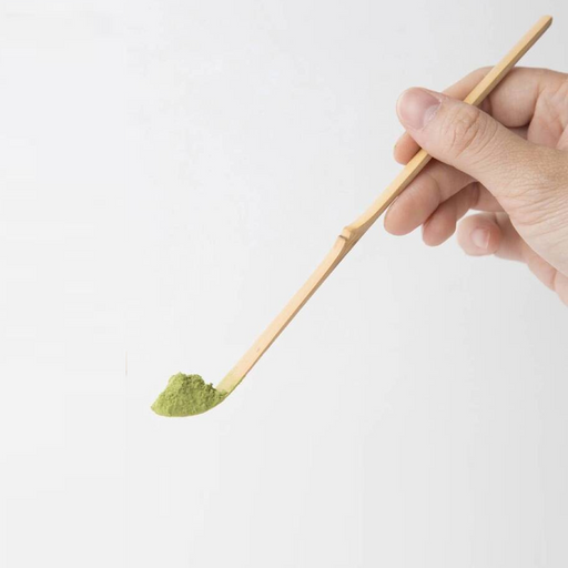 A hand holding a traditional Japanese bamboo matcha spoon (chashaku) with a scoop of green matcha powder on the tip.