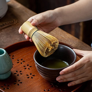 A hand holding a traditional Japanese Chasen bamboo matcha whisk over a dark ceramic bowl filled with freshly whisked green tea, placed on a wooden tray with a tea ceremony setup.